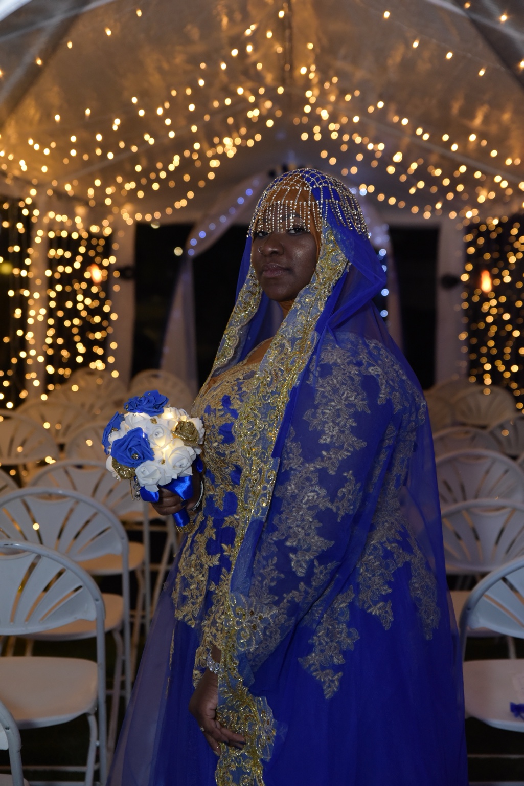 Bride in blue and gold holding a bouquet beneath warm string lights